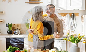 Happy senior elderly couple husband and wife embracing and dancing while cooking together in kitchen