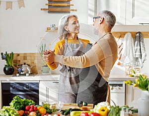 Happy senior elderly couple husband and wife embracing and dancing while cooking together in kitchen