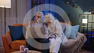 Happy senior elderly couple using laptop pc talking together doing online shopping at home sofa