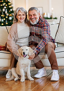 Happy senior family couple sit near decorated Christmas tree at home with dog golden retriever puppy