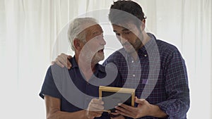 happy Senior Father and adult son having fun looking Picture frame remembering of the Past together on sofa in the room at home .