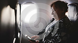 Happy senior female airplane passenger using smartphone messenger app, enjoying the view and smiling on the window seat.