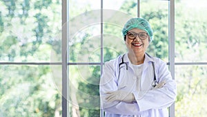 Happy senior female doctor in uniform and stethoscope standing near a window with crossed arms