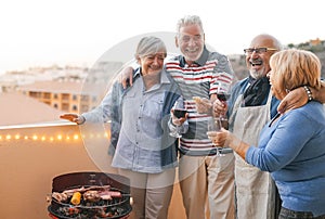 Happy senior friends having fun cheering with red wine at barbecue on terrace - Mature people dining and laughing together at bbq