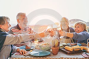 Happy senior friends having fun cheering with red wine at barbecue in terrace outdoor - Mature people making dinner