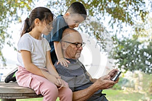 Happy senior grandfather using smart phone while grandson looks over his head, granddaughter sitting near.