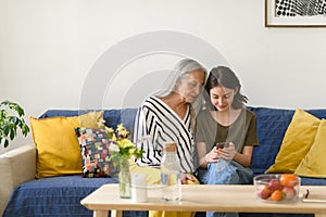 Happy senior grandmother with teenage granddaguhter sitting on sofa and using smartphone together at home.