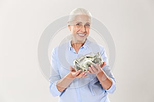 Happy senior lady in blue shirt holding glass bank with dollars, looking at camera