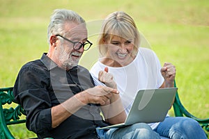 Happy senior loving couple relaxing with laptop computer at park excited together in morning time. old people sitting on a bench photo