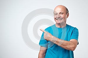 Happy senior man in blue tshirt looking at camera, smiling and pointing aside with hand.