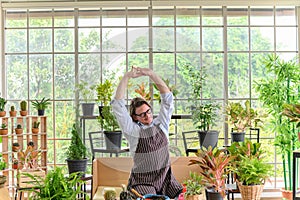 Happy senior man gardening wearing glasses and denim apron sitting in home gardening and stretching his arms, pulls hands up
