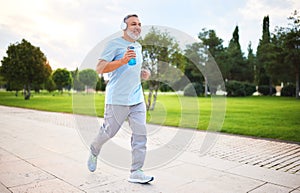 Happy senior man in headphones listening music while jogging outside in city park