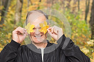 Happy senior man Holding and covering her eyes with two yellow maple leaves in the autumn city park. Active seniors.