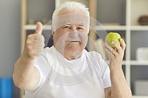 Happy senior man looking at camera holding fresh green apple and giving thumbs-up