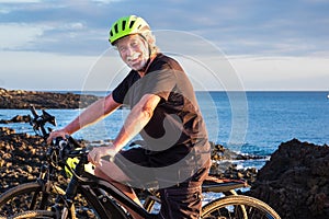 Happy senior man standing on the cliff riding his bici and looking at camera.Two electric bicycle close to him. Blue ocean water photo