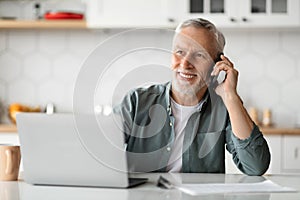 Happy Senior Man Talking On Cellphone And Using Laptop In Kitchen