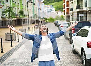 Happy senior man thanks God standing outdoors with hands spread wide open wearing jeans shirt with white t-shirt under