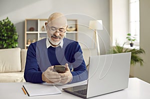 Happy senior man using his mobile phone while sitting at desk with laptop computer