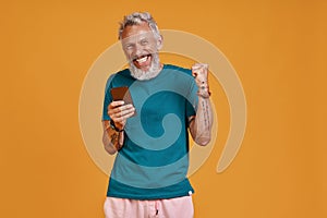 Happy senior man using smart phone and smiling while standing against orange background