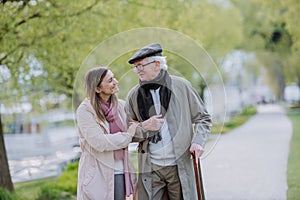 Happy senior man with walking stick and adult daughter outdoors on a walk in park.