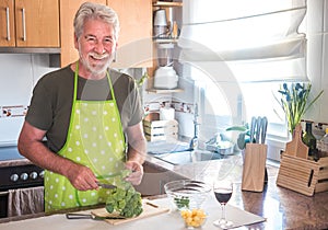 Happy senior man with white beard and hair in the kitchen while cuts and cleans the broccoli to prepare an healthy soup