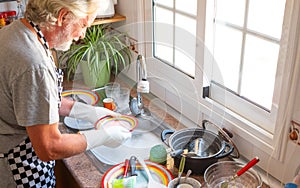 Happy senior man white hair and beard wearing apron washing dishes. Corner of kitchen. Grandfather at work. Bright light from