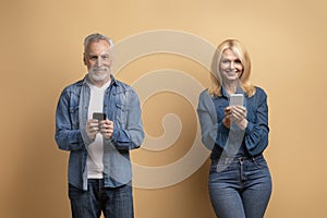 Happy senior man and woman using cell phones, beige background