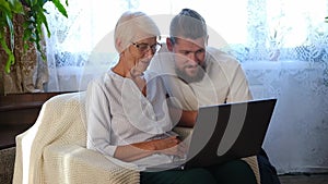 Happy senior mother, adult son sitting in living room at sofa and using laptop trying to make online payment. retirement
