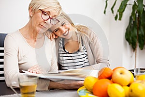Happy senior mother and her daughter looking at family photo album while sitting at a dining table. Daughter`s head resting on mot