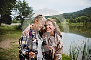 Happy senior mother hiker embracing with adult daughter by lake outdoors in nature