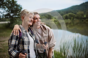 Happy senior mother hiker embracing with adult daughter when looking at lake outdoors in nature