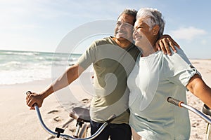 Happy senior multiracial couple with bicycles looking away at beach against sky on sunny day