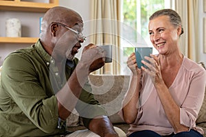 Happy senior multiracial couple having coffee while sitting on sofa at home