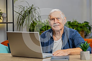 Happy senior old man grandfather sitting on sofa at table working on laptop pc at office home room