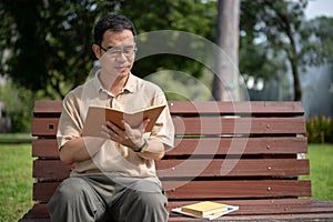 A happy senior retired Asian man is reading a book while relaxing on a bench in a green park