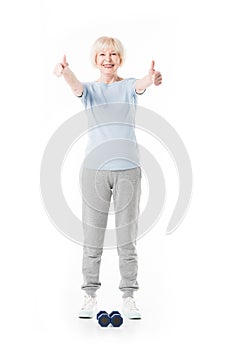 Happy senior sportswoman showing thumbs up gesture and dumbbells on floor