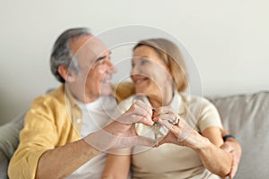 Happy senior spouses embracing and showing keys, making heart sign with hands, sitting ion sofa in living room interior