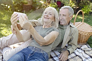 Happy senior spouses video calling on smartphone while having picnic and resting in garden at warm spring day