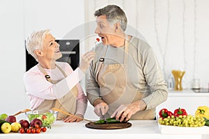 Happy Senior Wife Feeding Husband Cooking Together Standing In Kitchen