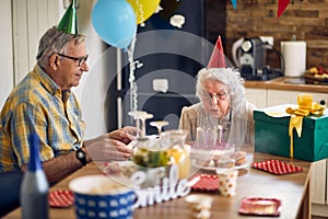 Happy senior woman blowing candles on her birthday cake, sitting at table with husband cheering her