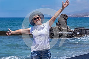 Happy senior woman with blue hat standing in front to the sea with open arms enjoying freedom and vacations