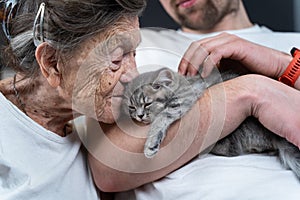 Happy senior woman cuddling and kiss, snuggle up to face small cute gray kitten, which held in arms by his grandson during visit