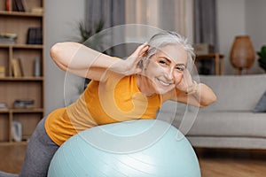 Happy senior woman doing exercises with fitness ball at home, training her back muscles during domestic workout