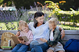 Happy senior woman with grandchildren sitting on bench and helping with homework outdoors in park.