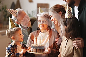 Happy senior woman holding cake with burning candles while celebrating birthday with big lovely family
