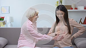 Happy senior woman holding pregnant granddaughters tummy, family replenishment