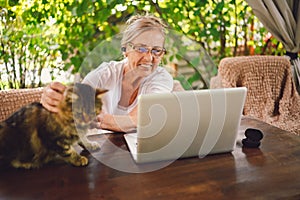 Happy senior woman with home cat use wireless headphones working online with laptop computer outdoor