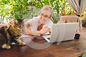 Happy senior woman with home cat use wireless headphones working online with laptop computer outdoor