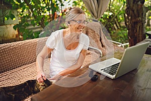 Happy senior woman with home cat use wireless headphones working online with laptop computer outdoor