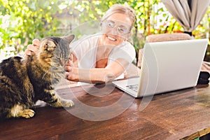 Happy senior woman with home cat use wireless headphones working online with laptop computer outdoor